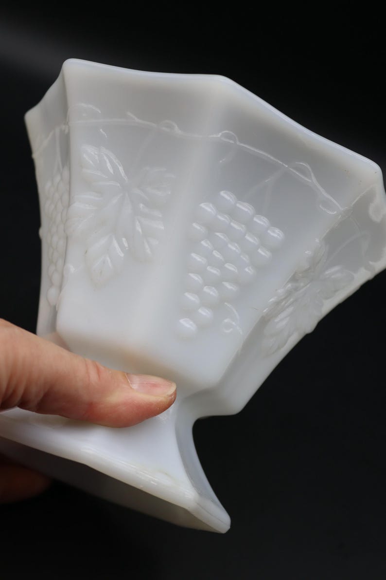 May include: A white, vintage-style glass bowl with a pedestal base. The bowl features embossed grape and leaf designs on its sides. The bowl is held by a hand, showcasing its shape and details against a dark background.