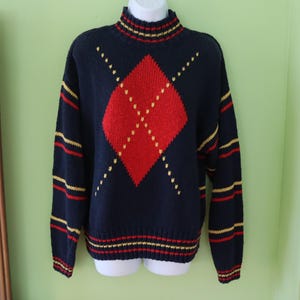 May include: A navy blue sweater with a red argyle pattern and yellow accents. The sweater has a mock turtleneck and striped details on the cuffs, hem, and collar. A classic, preppy style.