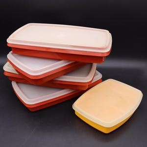 Puede incluir: Un conjunto de recipientes rectangulares para guardar alimentos, apilados. Las bases son naranjas y las tapas blanquecinas. Un recipiente tiene una base amarilla. Los recipientes son de plástico, probablemente vintage.