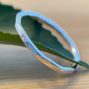 May include: A thin, silver-coloured ring with a hammered, faceted texture along the outer band. The metal surface is reflective and polished, catching the light to highlight the irregular, indented pattern that runs continuously around the circular band.