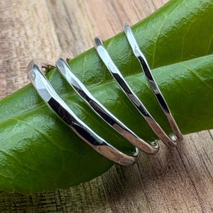 May include: A set of three silver-toned rings with a hammered, textured finish. The rings are arranged in a row, showcasing their reflective metallic surfaces and irregular, organic shapes. Each band features a polished sheen that catches the light.