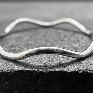 May include: A thin, polished silver ring featuring a continuous wavy or undulating design. The band has a smooth, reflective metallic surface that curves in a repeating pattern, creating a fluid shape that encircles the finger when worn.