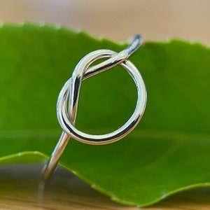 May include: A silver ring featuring a knot design. The ring's shiny metal reflects light, and it is set against a blurred green leaf. The knot design is the central element, offering a simple and elegant appearance.
