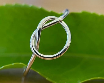 Op de afbeelding: Een gepolijste zilverkleurige ring met een enkel knoopontwerp. De band is glad en reflecterend en vormt een doorlopende lus die zichzelf kruist om een centraal knoopdetail te creëren. Het metalen oppervlak heeft een heldere uitstraling.