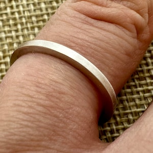 Puede incluir: Un anillo de metal plateado, fino y sencillo, con un acabado mate liso y un perfil de banda plano y uniforme. El anillo presenta un diseño limpio y sin adornos, sin piedras preciosas ni grabados, mostrando una textura metálica constante.