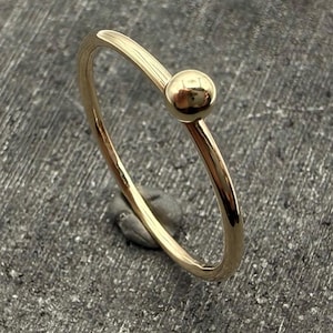 May include: A thin, polished gold ring featuring a single, smooth, spherical gold bead accent. The band is slender and circular, with a high-shine metallic finish that reflects light across its surface, creating a clean and simple jewellery piece.