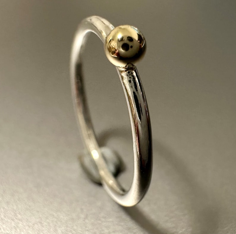 Peut inclure: Une fine bague en argent poli dot&eacute;e d'une perle dor&eacute;e sph&eacute;rique et lisse fix&eacute;e sur le dessus. Les surfaces m&eacute;talliques sont r&eacute;fl&eacute;chissantes, r&eacute;v&eacute;lant des reflets et des ombres qui soulignent la forme arrondie de l'anneau et de la perle.