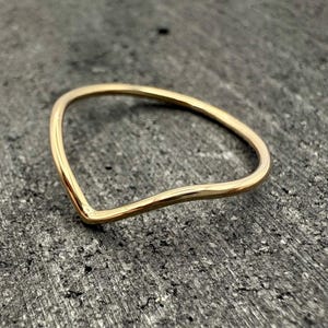 Op de afbeelding: Een dunne, gepolijste gouden ring met een gladde, doorlopende band die aan de voorzijde een opvallende V-vorm heeft. Het metaal heeft een reflecterende, warme afwerking en het ontwerp is eenvoudig, bestaande uit een enkele, ononderbroken gouden lus.