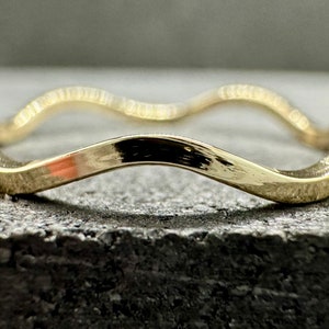 Peut inclure: Une bague en or poli présentant un design en forme de bande ondulée ou serpentine continue. La surface métallique est lisse et réfléchissante, captant la lumière le long des bords incurvés de la bague, qui conserve une épaisseur constante.