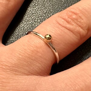 Peut inclure: Une fine bague en argent dot&eacute;e d'une unique petite perle sph&eacute;rique polie de couleur dor&eacute;e centr&eacute;e sur le dessus. La bague pr&eacute;sente une finition m&eacute;tallique lisse et r&eacute;fl&eacute;chissante ainsi qu'un design circulaire simple pos&eacute; sur le doigt.