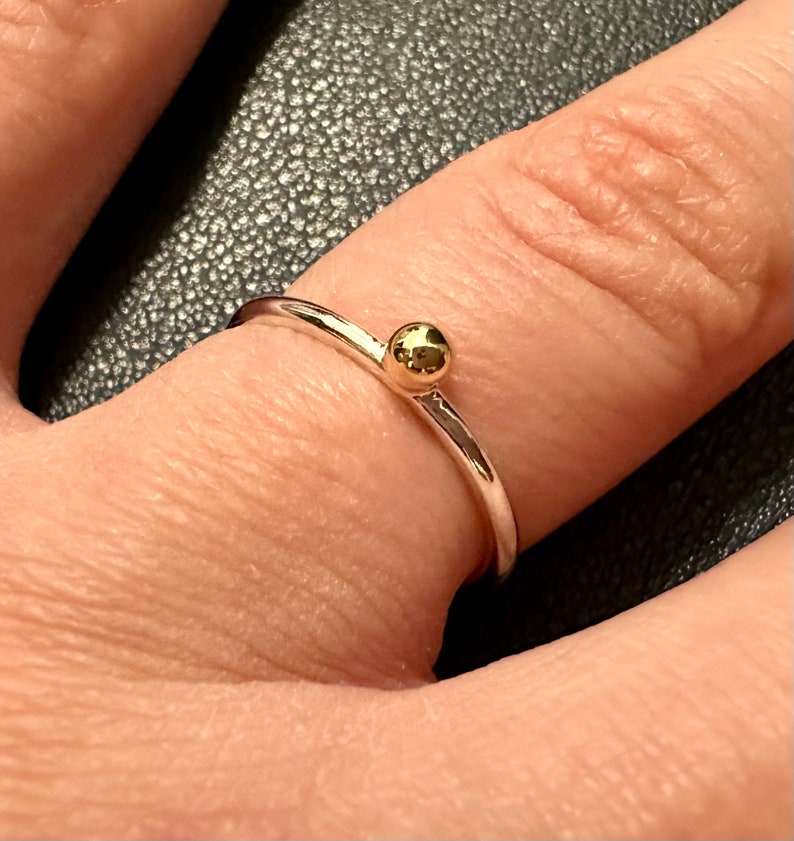 Peut inclure: Une fine bague en argent dot&eacute;e d'une unique petite perle sph&eacute;rique polie de couleur dor&eacute;e centr&eacute;e sur le dessus. La bague pr&eacute;sente une finition m&eacute;tallique lisse et r&eacute;fl&eacute;chissante ainsi qu'un design circulaire simple pos&eacute; sur le doigt.