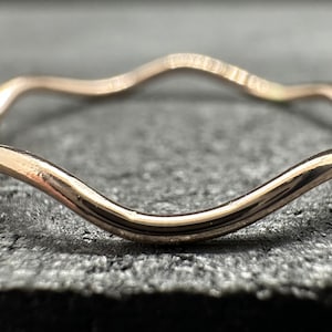 May include: A thin, polished rose gold ring featuring a continuous, gentle wavy or undulating design. The metal surface is smooth and reflective, showcasing a soft pinkish-gold hue that curves consistently around the entire circumference of the band.