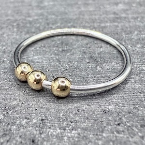 May include: A silver ring with three gold-colored beads. The ring has a thin band and the beads are evenly spaced across the top. The ring is set against a textured gray background.