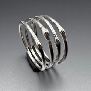 Peut inclure: Un ensemble de quatre bracelets argentés. Chaque bracelet a un design lisse et arrondi avec un centre légèrement bulbeux. Les bracelets sont empilés, créant un effet de superposition. Les bijoux sont présentés sur une surface sombre sur fond gris.