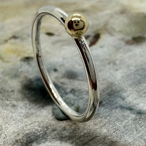 Peut inclure: Une fine bague en argent poli dot&eacute;e d'une unique petite perle sph&eacute;rique lisse de couleur dor&eacute;e fix&eacute;e sur le dessus. La bague pr&eacute;sente une finition m&eacute;tallique r&eacute;fl&eacute;chissante et un design circulaire simple d'une &eacute;paisseur constante.