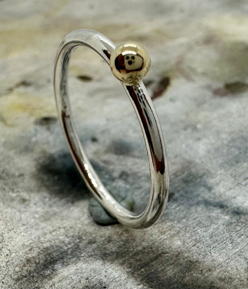 Peut inclure: Une fine bague en argent poli dot&eacute;e d'une unique petite perle sph&eacute;rique lisse de couleur dor&eacute;e fix&eacute;e sur le dessus. La bague pr&eacute;sente une finition m&eacute;tallique r&eacute;fl&eacute;chissante et un design circulaire simple d'une &eacute;paisseur constante.