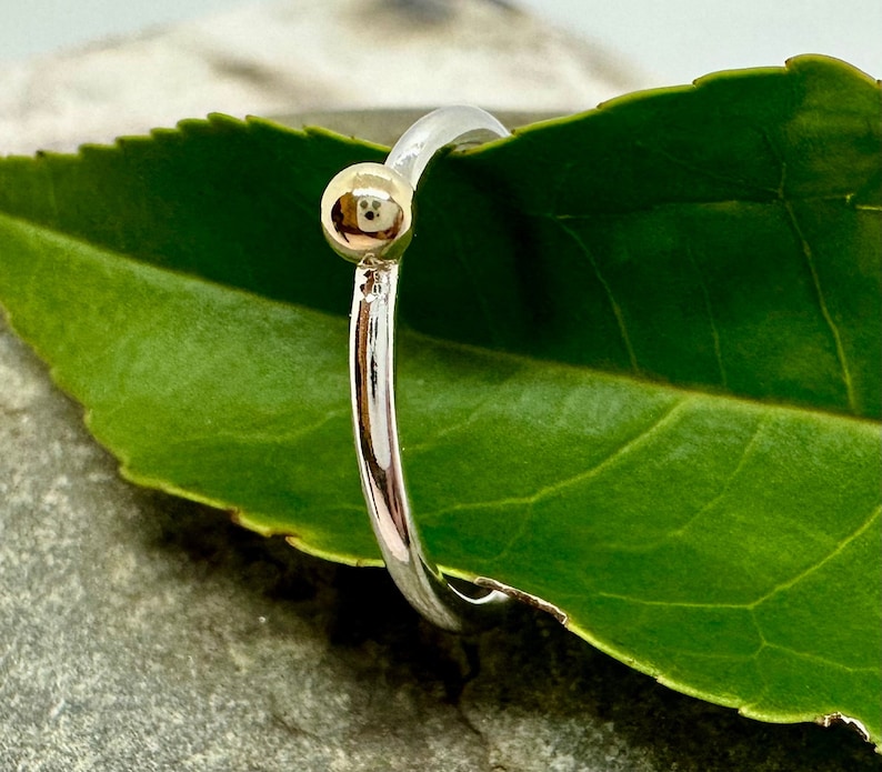 Peut inclure: Une fine bague en argent poli orn&eacute;e d'une unique petite perle sph&eacute;rique de couleur dor&eacute;e. L'anneau est circulaire et r&eacute;fl&eacute;chissant, pr&eacute;sentant un &eacute;clat m&eacute;tallique qui contraste avec le design simple et &eacute;pur&eacute; de la perle.