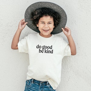May include: A young person wearing a white t-shirt with the text "do good be kind" printed on it. They are wearing a gray hat and blue jeans.
