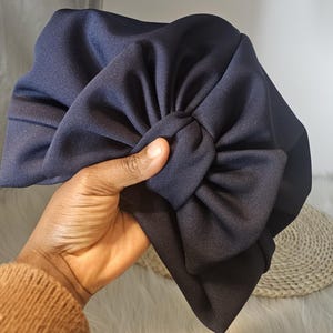 May include: A navy blue turban with a large bow. The head covering is made of a smooth fabric. The bow is a prominent feature, positioned centrally on the front of the turban.