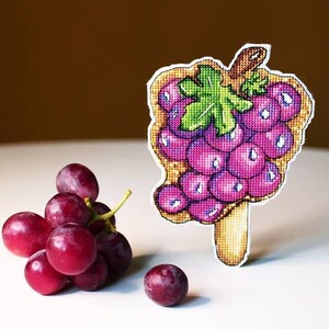 May include: A cross-stitch embroidery of a bunch of purple grapes with green leaves and a brown stem. The grapes are surrounded by a white outline. There is a small bunch of real grapes and a single grape on a white surface.