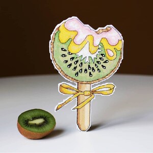 May include: A cross-stitch pattern of a kiwi fruit on a stick with a yellow bow. The kiwi fruit is green with black seeds and has a pink and yellow topping. A whole kiwi fruit is in the foreground.