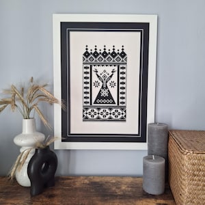 May include: Black and white framed embroidery depicting a stylized figure with outstretched arms, surrounded by geometric patterns.