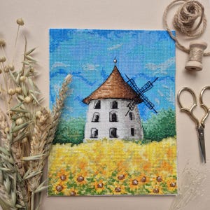 May include: A cross-stitch embroidery of a white windmill with a brown roof, surrounded by a field of yellow sunflowers against a blue sky.