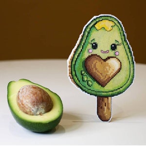 May include: A green avocado popsicle with a heart and a smiling face. A halved avocado with a seed is in the background.