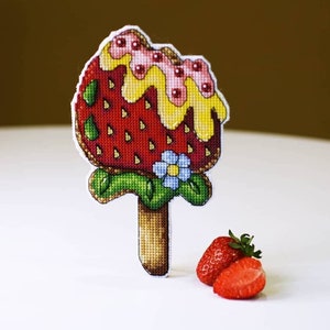 May include: A cross-stitch embroidery of a strawberry popsicle with a yellow and pink topping. The popsicle is on a stick and has green leaves. There are two real strawberries in the foreground.