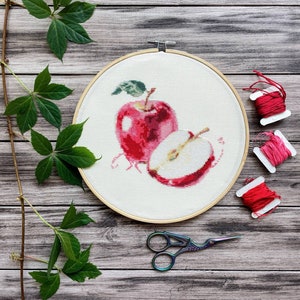 May include: A cross-stitch embroidery of a red apple and a sliced apple half, set in a wooden hoop. The embroidery is on white fabric. Red and pink thread spools and small scissors are next to the hoop. Green leaves and vines are in the background.
