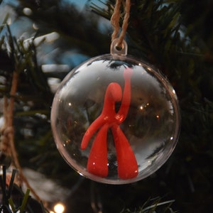 Op de afbeelding: Een heldere, ronde kerstbal met een rode abstracte figuur erin, hangend aan een touwtje. De bal hangt tegen een achtergrond van groene kerstboomtakken en lichtjes.