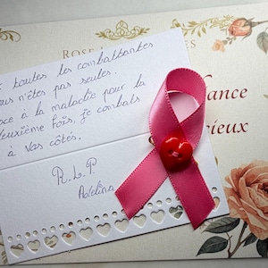 May include: A white card with a scalloped heart-shaped edge featuring handwritten French text and a pink ribbon pinned with a small red heart-shaped button. The card rests on a larger rectangular card decorated with vintage-style rose illustrations.