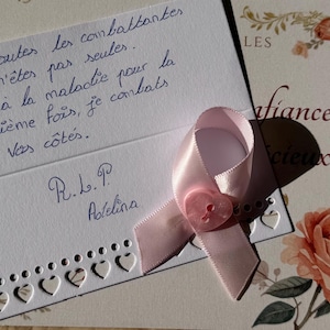 May include: A white card with a decorative heart-shaped border and handwritten French text, featuring a light pink satin ribbon pinned with a small, heart-shaped pink button. The card includes the text: Pour toutes les combattantes, vous n'êtes pas seules. Face à la maladie pour la deuxième fois, je combats à vos côtés. R.L.P. Adelina.