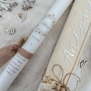 May include: A white candle with floral accents, the name "Antonia", and the date 01.03.2026 printed on it. A wooden box with the name "Anton" in white script is next to the candle. Decorative wooden accents and a ribbon are also present.