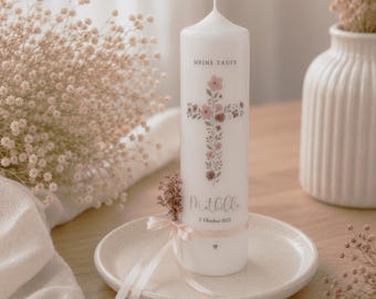 May include: A white cylindrical pillar candle featuring a floral cross design in shades of pink and brown. The text MEINE TAUFE, the name Mathilda, and the date 2. Oktober 2025 are printed on the front. A small pink ribbon and dried flowers are attached.