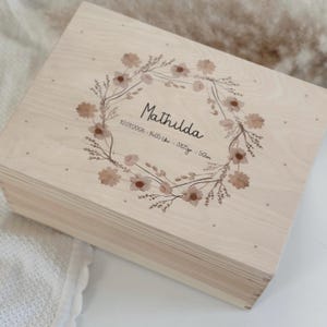 Könnte beinhalten: Eine hellfarbene Holzkiste mit einem floralen Kranzdesign. Der Kranz umgibt den Namen "Mathilda" und enthält Geburtsdetails. Die Kiste ist quadratisch und hat eine natürliche Holzmaserung mit einer weichen, neutralen Farbpalette.