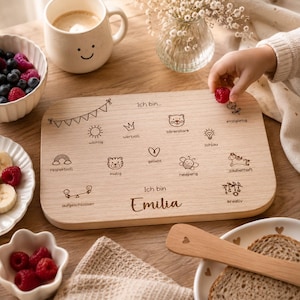 May include: A rectangular wooden board featuring laser-engraved illustrations and German text including the name Emilia. The board displays icons like a sun, bear, heart, and bee, each paired with descriptive words, alongside a bunting banner design.