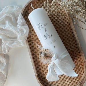 May include: White pillar candle with the names "Elisa & Max" and the date "11 Juli 2024" written on it. The candle is decorated with a white ribbon and dried flowers.