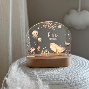 Puede incluir: Una luz nocturna de acrílico en forma de arco con base de madera que presenta una escena submarina con una ballena, medusas, una tortuga marina y un caballito de mar. El texto Elias y 12.11.2025 está impreso en blanco sobre la superficie.