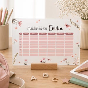 Könnte beinhalten: Ein rechteckiges Wochenplan-Board aus Acryl mit der Aufschrift Stundenplan von Emilia, einem rosa Raster für acht Unterrichtsstunden und den Wochentagen von Montag bis Freitag, verziert mit Aquarellillustrationen von rosa Blumen und Schmetterlingen.