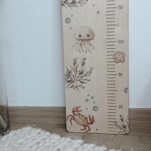 K&ouml;nnte beinhalten: Eine hellfarbene Holzwachstumsmesslatte mit Aquarellillustrationen von einer Qualle, einer Krabbe, Seetang und Muscheln. Das Brett enth&auml;lt eine vertikale Messskala mit Markierungen von 80 bis 110 Zentimetern in schwarzer Druckfarbe.