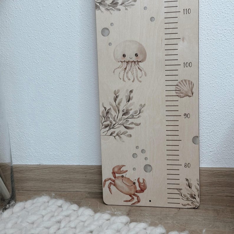 K&ouml;nnte beinhalten: Eine hellfarbene Holzwachstumsmesslatte mit Aquarellillustrationen von einer Qualle, einer Krabbe, Seetang und Muscheln. Das Brett enth&auml;lt eine vertikale Messskala mit Markierungen von 80 bis 110 Zentimetern in schwarzer Druckfarbe.