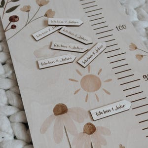 K&ouml;nnte beinhalten: Eine helle Holzwachstumsmesslatte mit Blumenillustrationen, einer Sonnengrafik und einer Skala bis 100. Enthalten sind mehrere kleine, pfeilf&ouml;rmige Holzmarker, die jeweils mit deutschem Text bedruckt sind und das Alter von 1 bis 9 Jahren angeben.