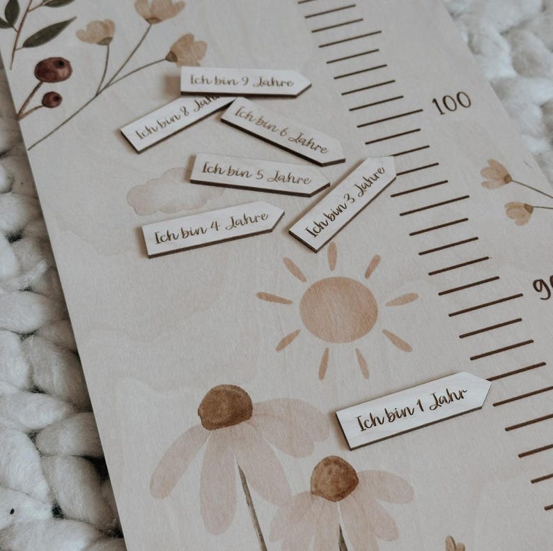 K&ouml;nnte beinhalten: Eine helle Holzwachstumsmesslatte mit Blumenillustrationen, einer Sonnengrafik und einer Skala bis 100. Enthalten sind mehrere kleine, pfeilf&ouml;rmige Holzmarker, die jeweils mit deutschem Text bedruckt sind und das Alter von 1 bis 9 Jahren angeben.