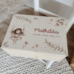 Può includere: Scatola ricordo in legno chiaro con illustrazione di una bambina su un'altalena, decorazioni floreali e il nome "Mathilda". La scatola include i dettagli della nascita: data, ora, peso (3120g) e lunghezza (52cm).