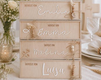 May include: A stack of four rectangular light-coloured wooden boxes, each featuring the text Taufkerze von followed by a white script name: Emilia, Emma, Malina, or Luisa. Each box is decorated with twine, small wooden beads, and dried flowers.