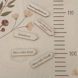 K&ouml;nnte beinhalten: Eine helle Holzwachstumsmesslatte mit Blumenmuster und einer vertikalen Skala von 100 bis 110. F&uuml;nf pfeilf&ouml;rmige Holzmarker sind enthalten mit den deutschen Texten: Kindergartenkind, Ersten Milchzahn verloren, Schulkind, Mein erstes Wort und Mein erster Schritt.