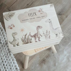 May include: A light wooden box with a hinged lid, decorated with watercolor illustrations of dinosaurs, foliage, and a sun. The lid features the name "FELIX" and birth details. The box is placed on a small wooden stool.