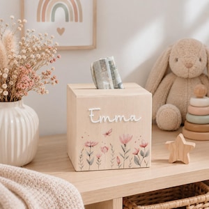 Può includere: Una scatola quadrata in legno chiaro con una fessura sulla parte superiore, caratterizzata dal nome Emma in corsivo bianco e da un disegno stampato di fiori rosa e una piccola farfalla. La scatola è posizionata su una superficie in legno.