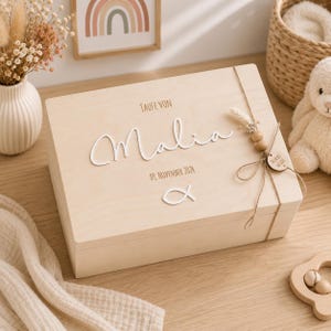 May include: A rectangular light-colored wooden keepsake box featuring the engraved text "Taufe von Malia" and "09. November 2024" with a white 3D script name "Malia" and a white fish symbol. The box is decorated with twine, wooden beads, and a dried flower.